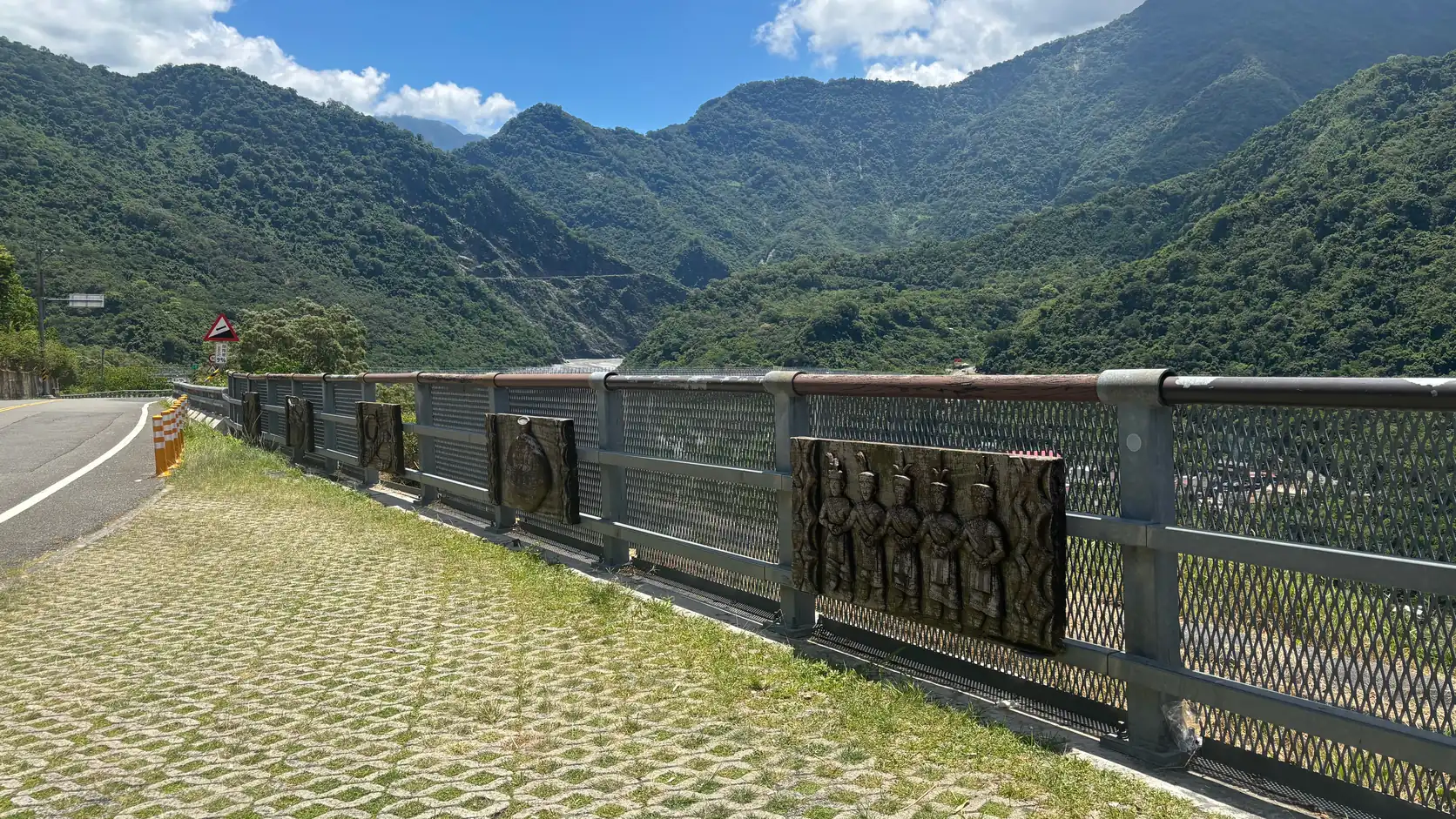 Wooden relief portraits of Indigenous people set into the railing of a bridge on a mountain road, with green ridges and a river valley in southern Taiwan beyond
